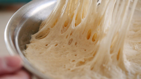 Dough in a bowl that is being stretched to show the formation of gluten